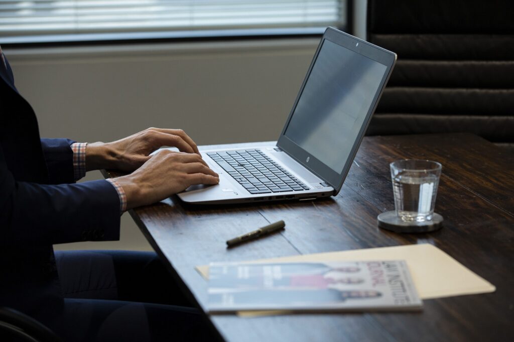 Person in a navy suit typing on a silver laptop at a dark wooden table beside a glass of water.