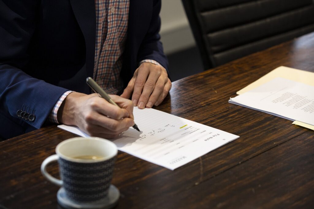 Person in a navy blazer writing with a pen on a printed form at a dark timber table.